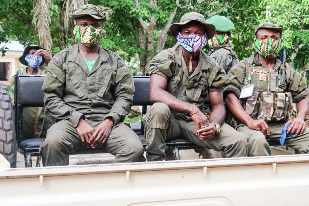 A group of soldiers wearing masks and olive-green uniforms ride in the back of a truck.