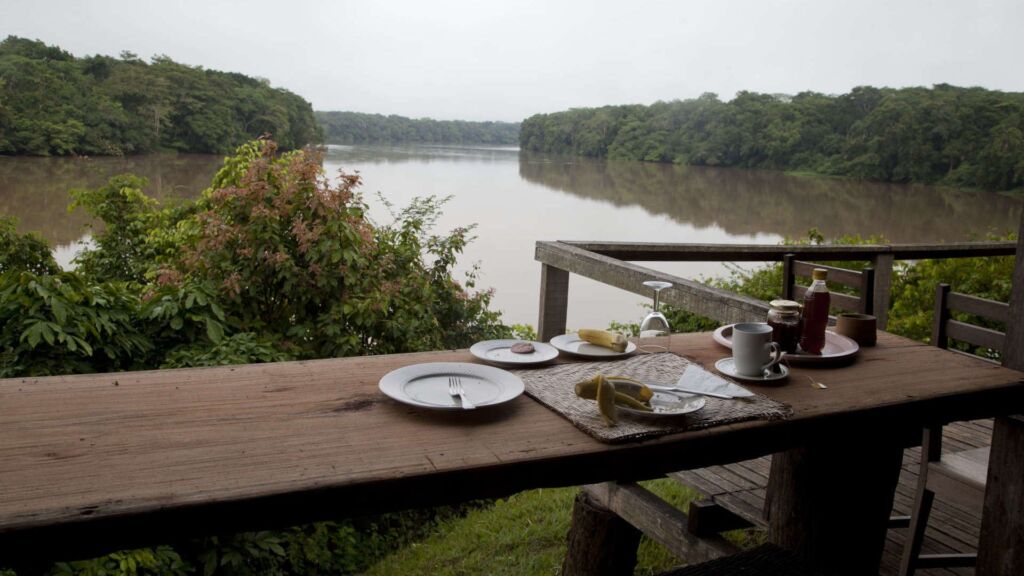 A breakfast table overlooking a wide, calm river lined with dense, green trees under a gray sky.