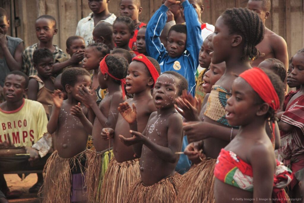 A group of African children wearing grass skirts and red headbands are clapping and singing in unison.