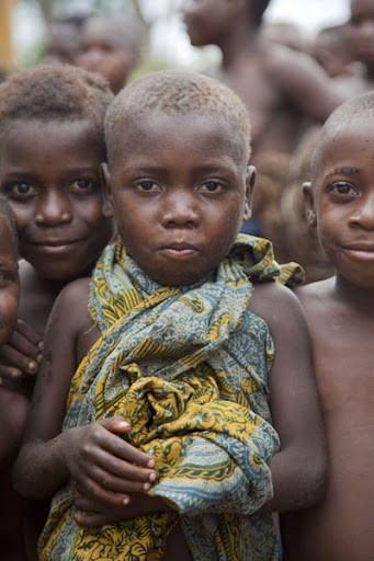 This photo shows a group of African children, with the focus on a child in the center wrapped in a patterned cloth, looking directly at the camera with a serious expression.