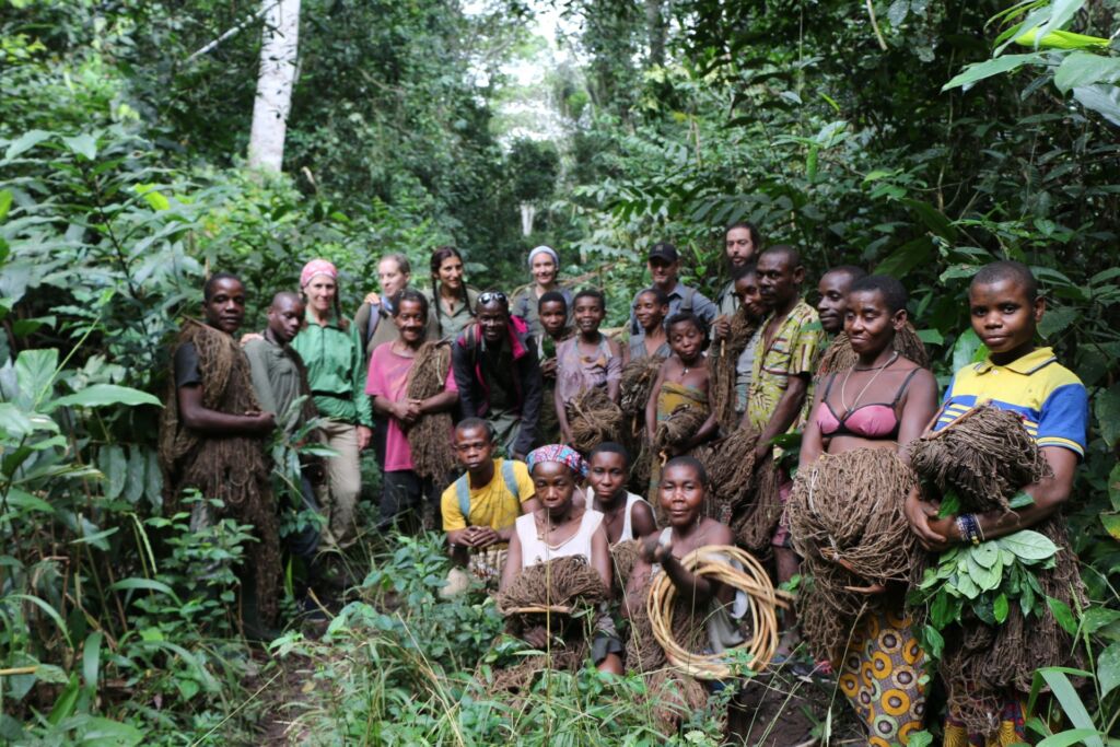 A large group of adults and children stand in a lush green forest, holding bundles of natural materials, with a mix of traditional clothing and Western attire.