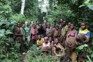 A large group of adults and children stand in a lush green forest, holding bundles of natural materials, with a mix of traditional clothing and Western attire.