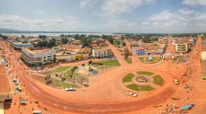 A high-angle panoramic shot depicts a bustling African town with reddish-brown roads, buildings with rusty roofs, and roundabouts with green spaces, hinting at a vibrant community.