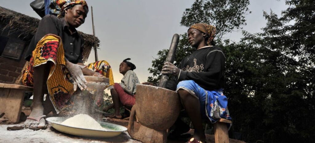 Three African women are preparing food using traditional methods, one grinding grain with a stone and mortar, one grinding it with her hands, and one sitting nearby.