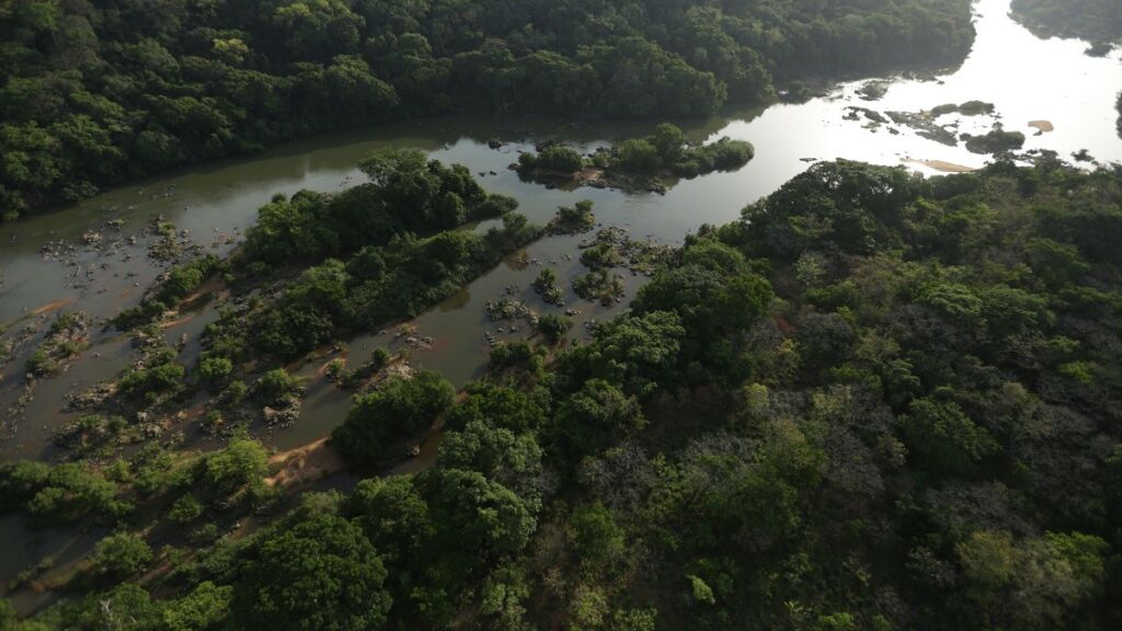 An aerial view shows a river winding through a dense, green forest with small islands and rocky areas visible in the water.