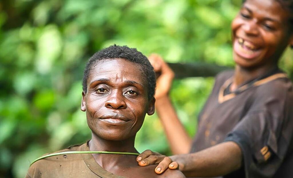 A woman with short hair and a small smile poses for a photo while another woman behind her laughs and touches her shoulder in a lush, green environment.