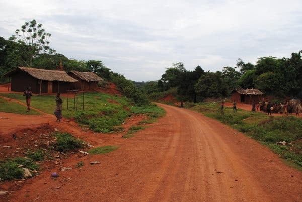 A red dirt road winds through a rural African village with simple huts and people gathered along the roadside under an overcast sky.