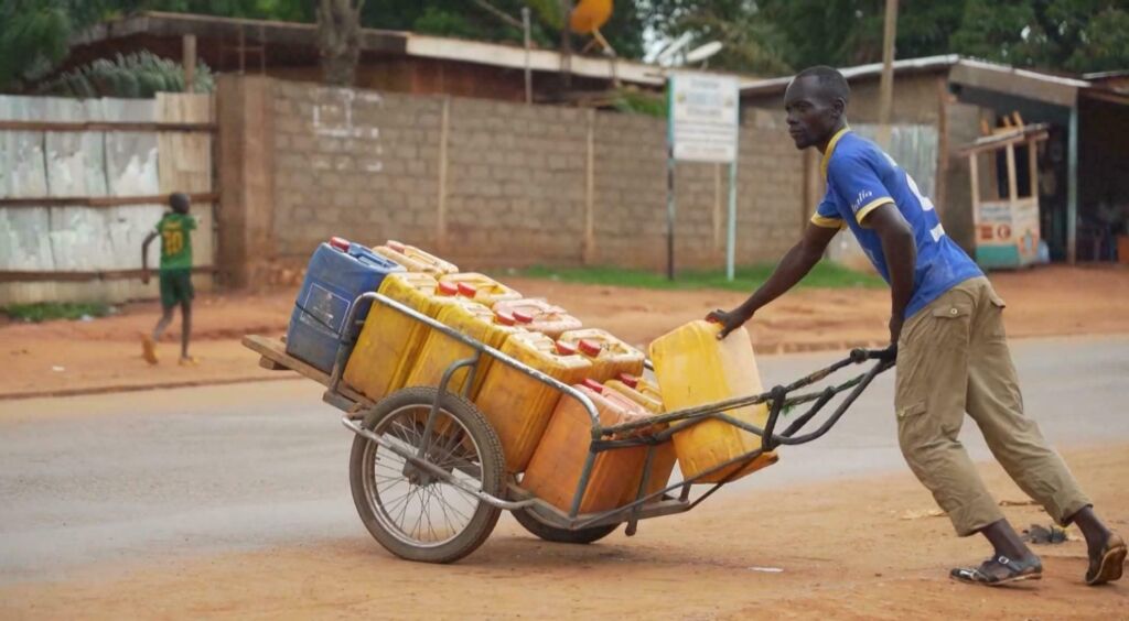 A man pulls a cart loaded with yellow jerrycans down a dirt road in an African village. A child in a green jersey runs in the background.