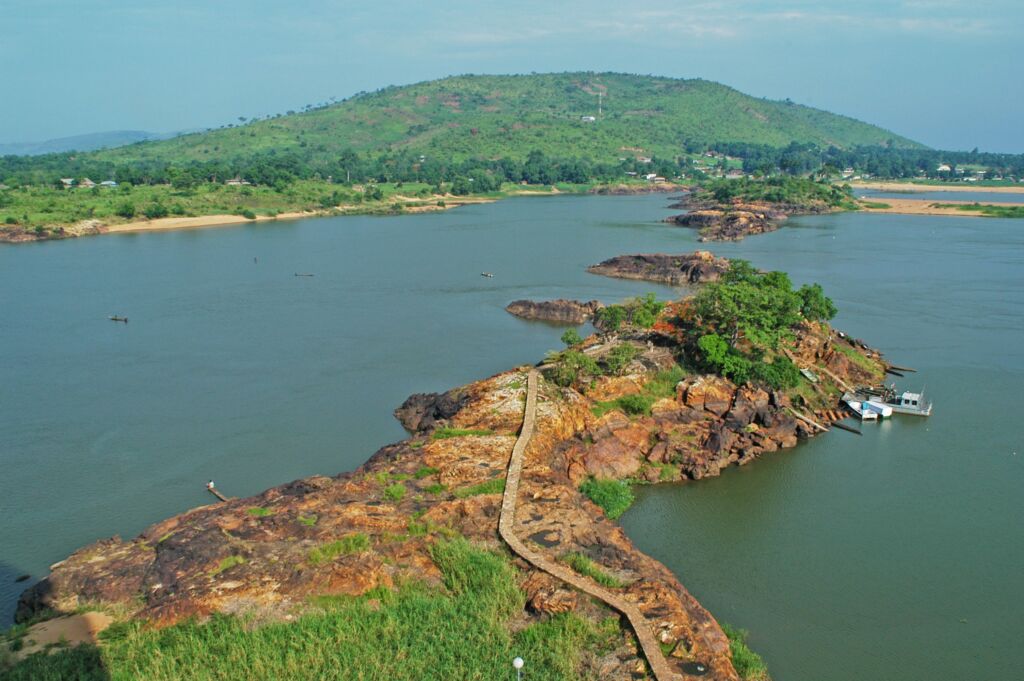 A winding stone path leads across rocky islands in a wide river, with a lush green hill in the background and boats docked at the furthest island.