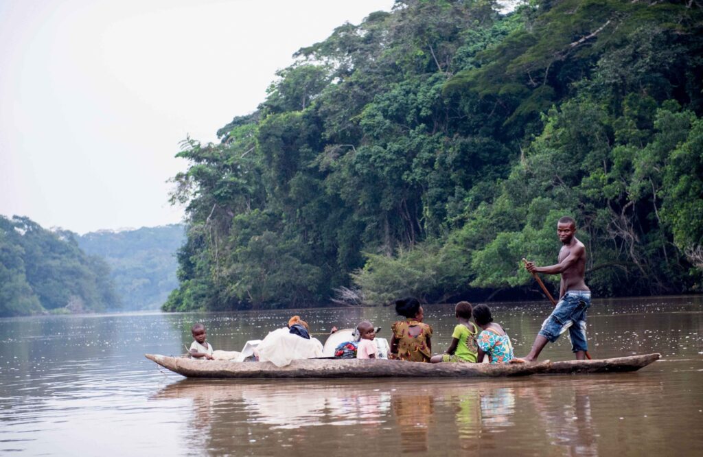 A family is traveling down a river in a dugout canoe, with a man standing and using a pole to propel the boat forward.