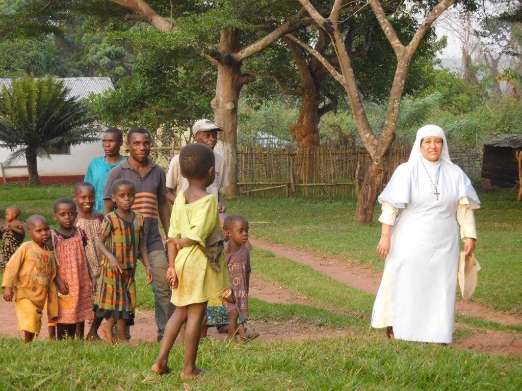 A nun in a white habit smiles at a group of African children and men standing on a grassy path in a rural area.