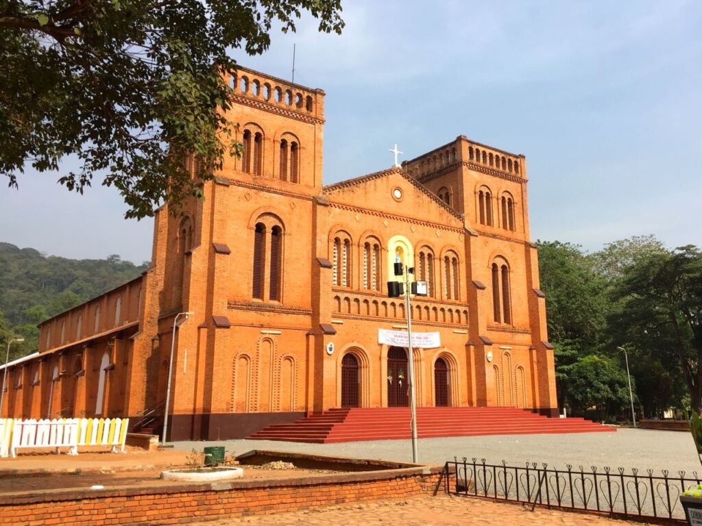 A large, red-brick church with two towers and a cross on top stands prominently in a landscape with trees and hills in the background.