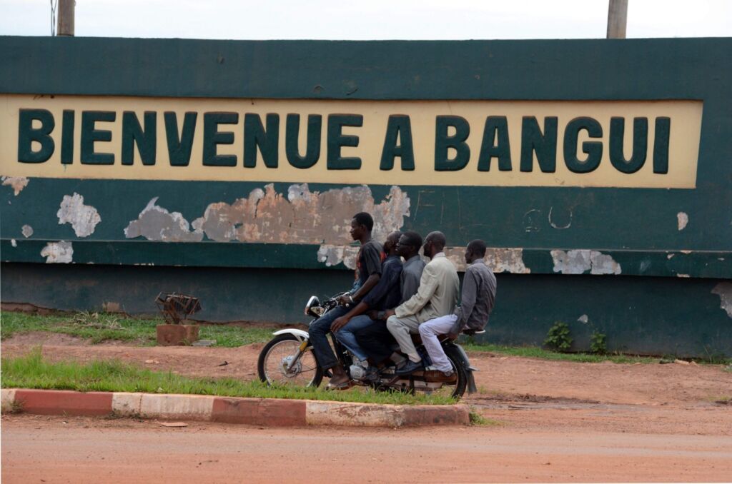 Five men ride a motorbike in front of a wall sign that reads