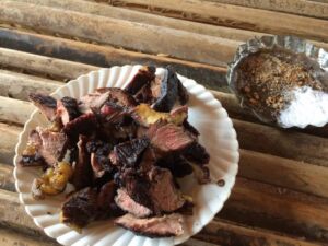 The image shows a white plate filled with chunks of grilled meat, alongside a small container with a dark sauce sprinkled with white and brown grains, all resting on a bamboo surface.