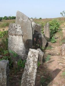 A cluster of ancient, weathered stone slabs stands amidst sparse vegetation and parched earth in a rural landscape.