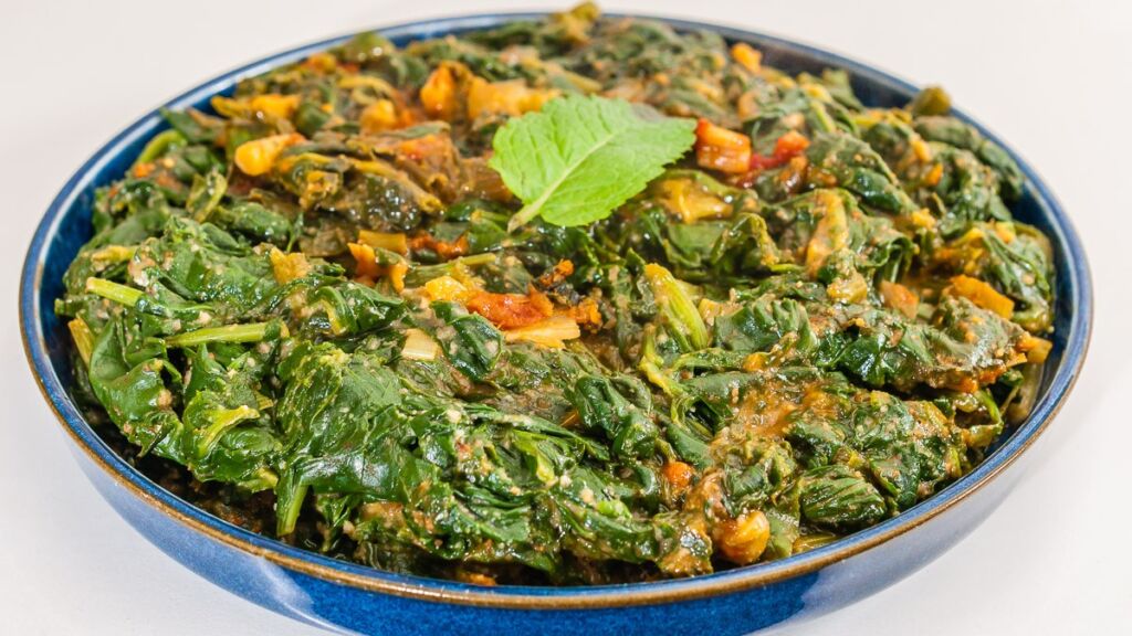 A blue bowl filled with cooked spinach, garnished with a mint leaf, is displayed on a white surface.