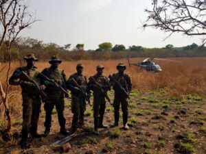 Five armed soldiers in camouflage stand guard in a field, with a helicopter visible in the background.