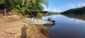 Boat Rental in Central African Republic A silver boat with a canopy is moored on a sandy riverbank under a blue sky with trees in the background.