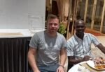 Two men are seated at a table, one in an Adidas t-shirt, the other in a striped shirt, with a plate of food between them.