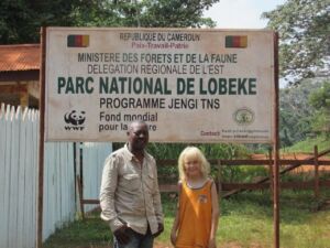 Two people are posing in front of a sign for Parc National de Lobeke in Cameroon, featuring text in French, WWF logo, and contact information.