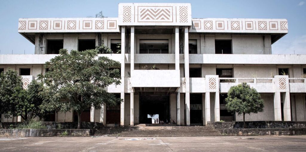 A weathered, multi-story building with geometric patterns stands overgrown with vegetation, its windows dark and stairs leading to a shadowed entrance where figures can be seen.