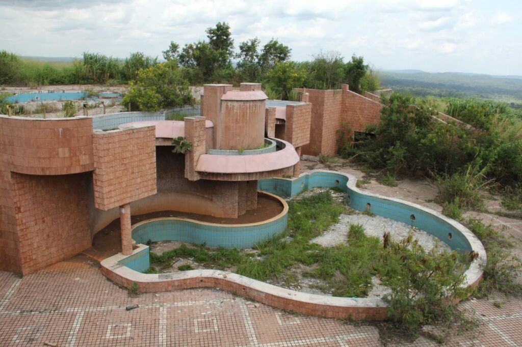 An abandoned and overgrown resort complex features a unique architectural design with multiple pools, brick structures, and a view of the surrounding landscape.