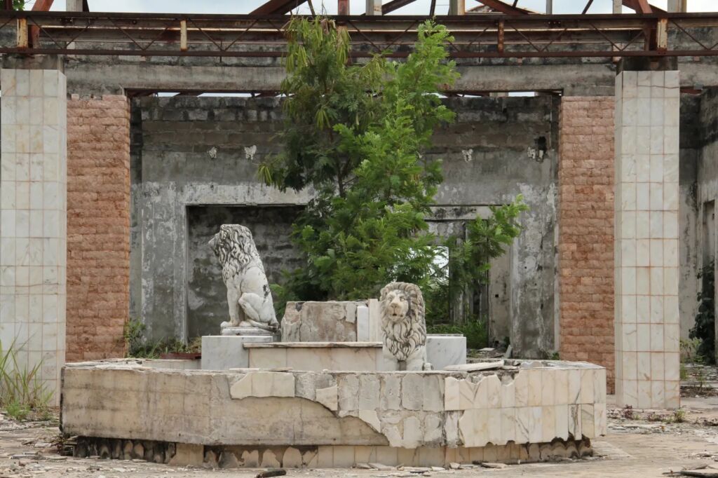 An abandoned building featuring two lion statues in the center, surrounded by weathered walls and overgrown vegetation.