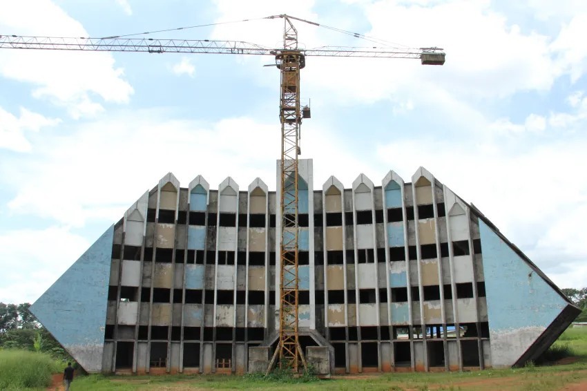 A construction crane stands tall in front of a large, unfinished building with a unique, angular design and blue accents.