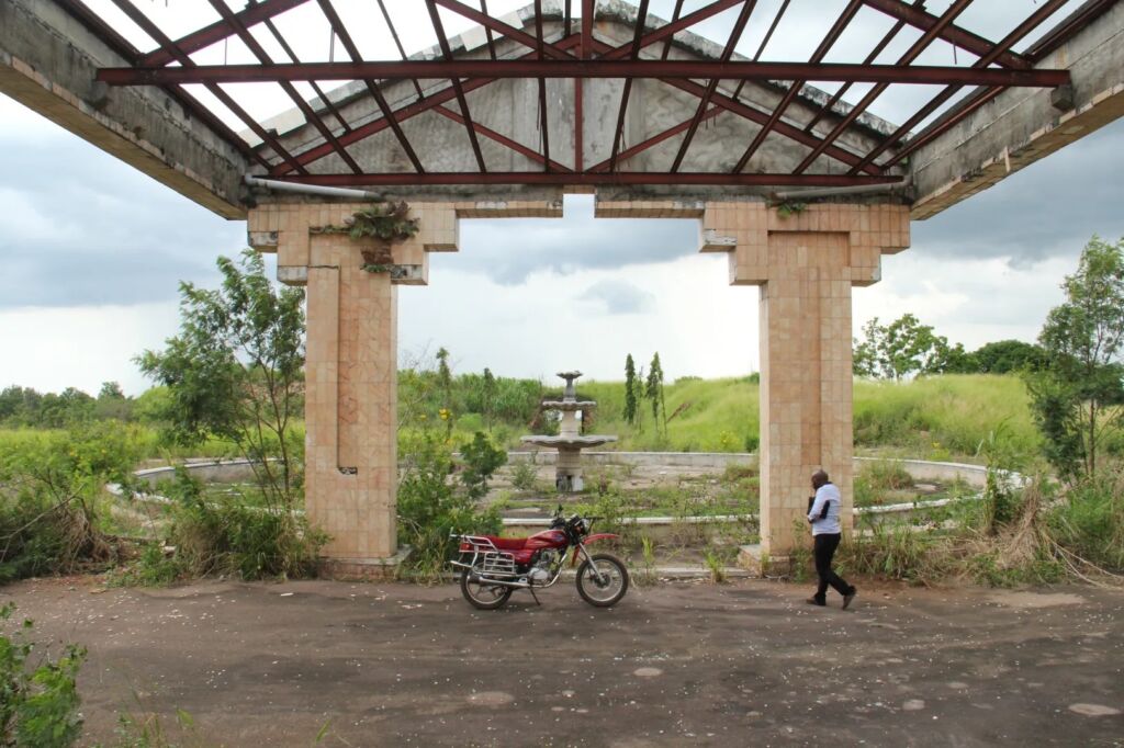 An overgrown area features a red motorcycle, a dilapidated building frame, and a man walking by, with a fountain visible in the background.