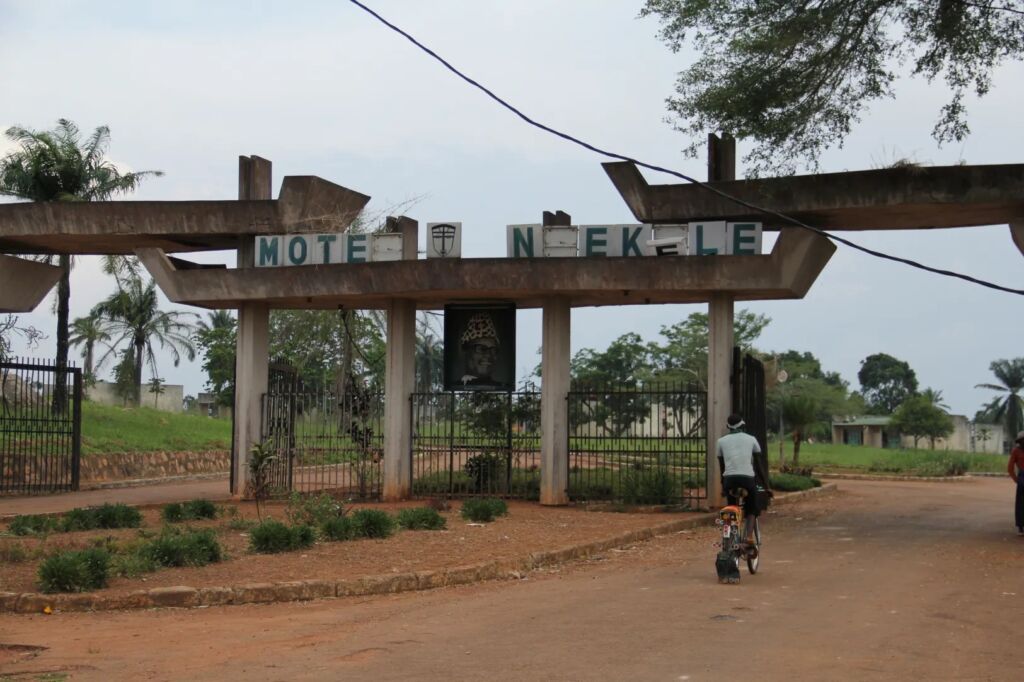 A person rides a bicycle through the entrance gate of Motel Nakele, featuring a large sign and a portrait.