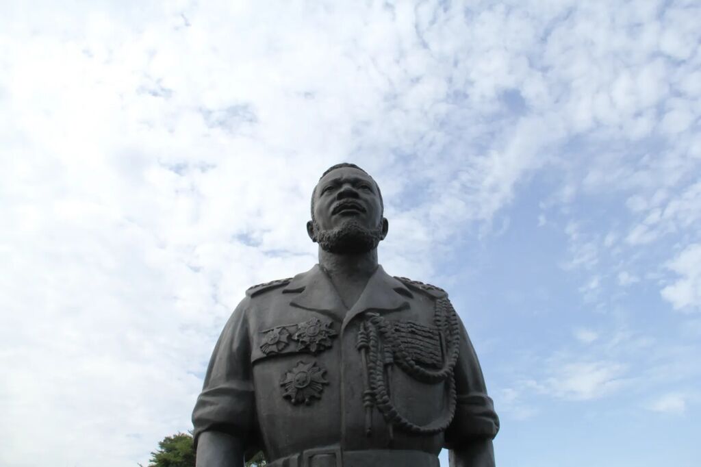 A low-angle shot captures a statue of a man in military attire against a bright, cloudy sky.