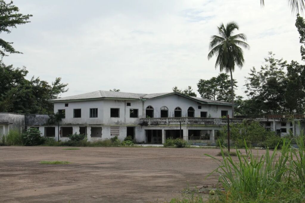 The image shows a large, two-story, abandoned white building with many open windows and a crumbling facade, surrounded by overgrown vegetation.