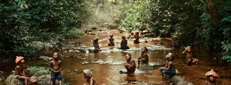 A group of children stand and sit in a stream surrounded by lush green vegetation, gazing upwards.