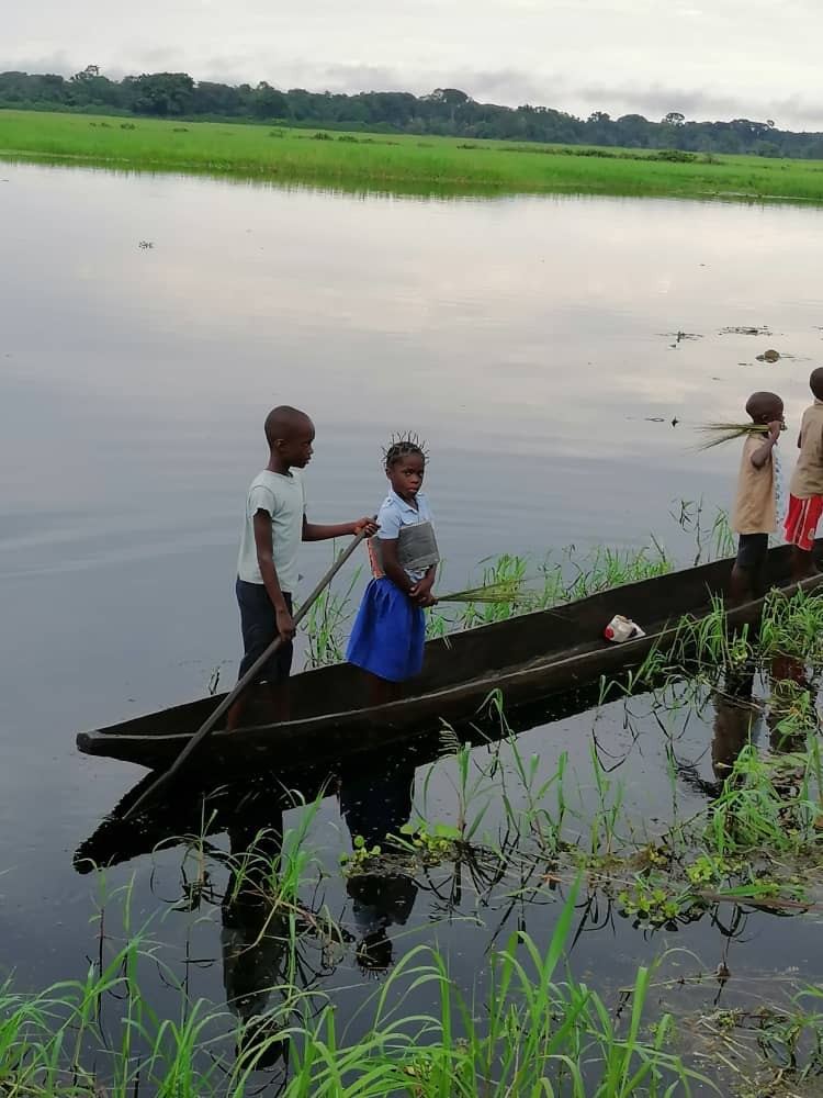 Children are on a wooden canoe navigating through a river with grassy shores, seemingly on a cloudy day.
