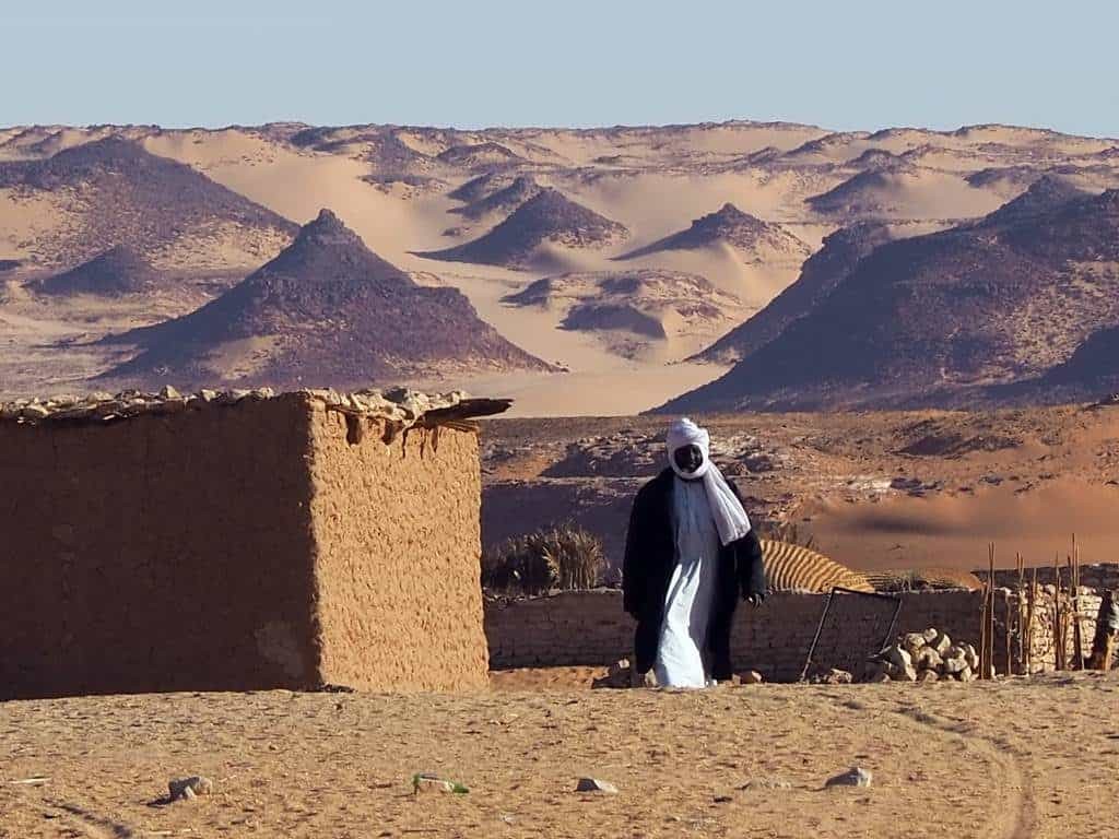A man in traditional dress stands in a desert landscape with mud buildings and pyramidal hills in the background.