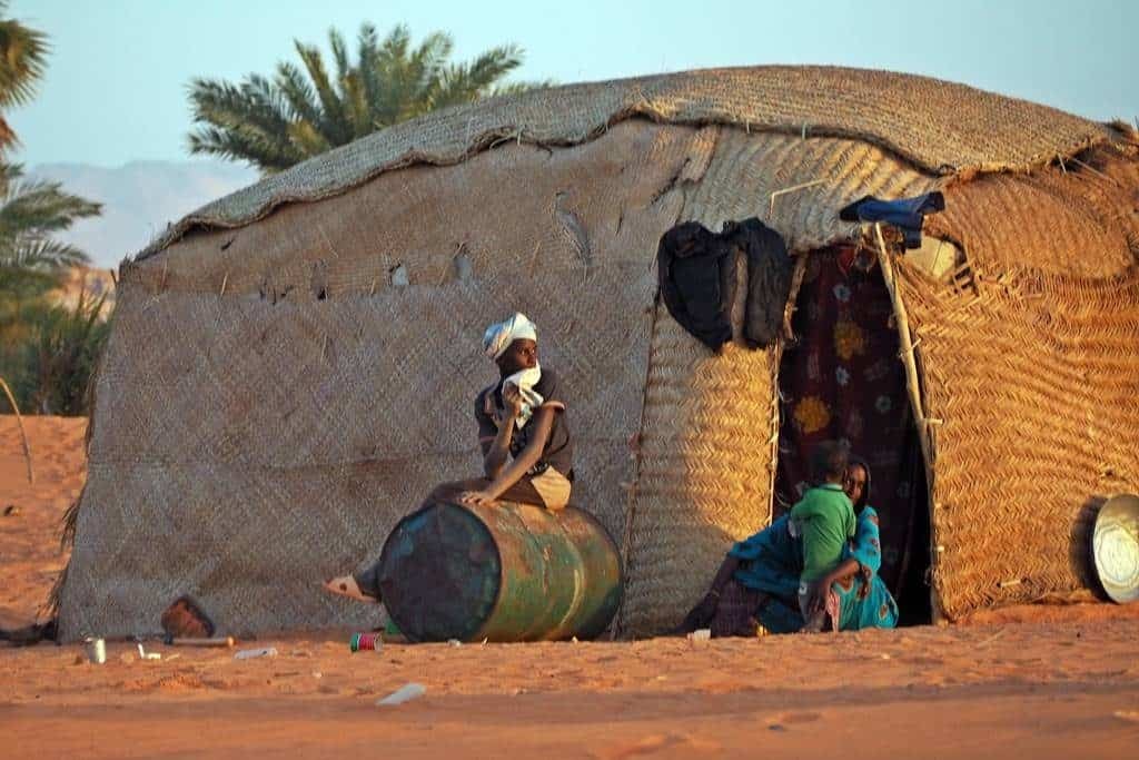 A man sits on a barrel outside a thatched hut, while a woman and child are inside, in a desert setting.