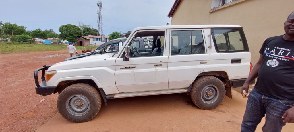 A white Toyota Land Cruiser stands on a red dirt surface, with a man wearing a black t-shirt standing beside it, under a cloudy sky.