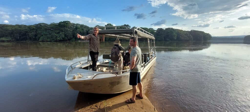 A group of people stands on a boat docked on the riverbank, surrounded by lush greenery and a partly cloudy sky.