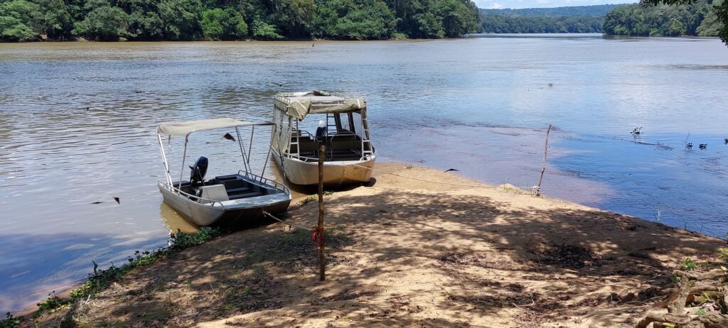 Two boats are docked on a sandy riverbank, with lush green trees lining the opposite shore and the river flowing calmly in the foreground.