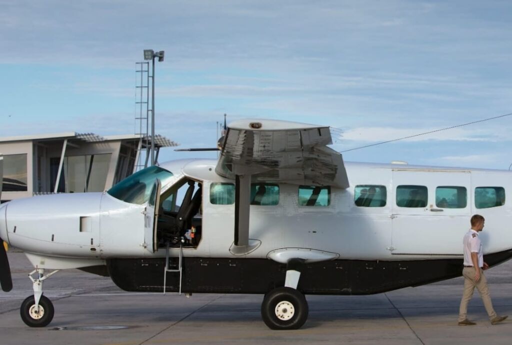A small white airplane with a pilot walking beside it sits on the tarmac at an airport.