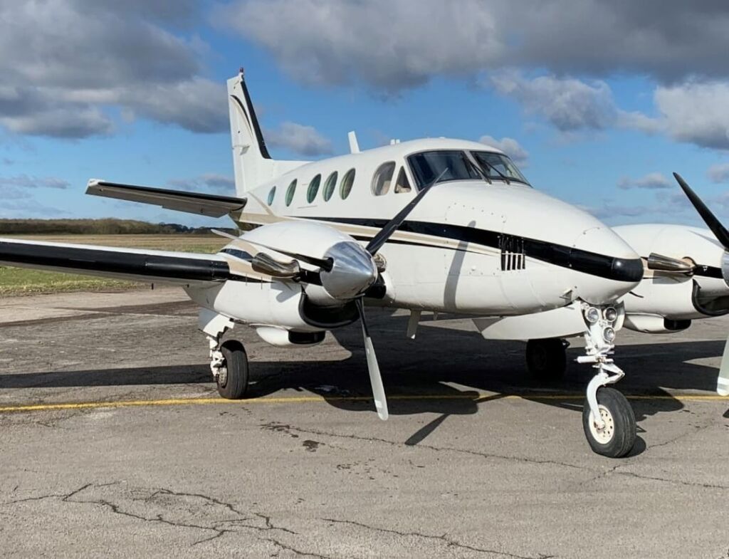 A white and black twin-engine turboprop airplane is parked on an airfield under a partly cloudy sky.
