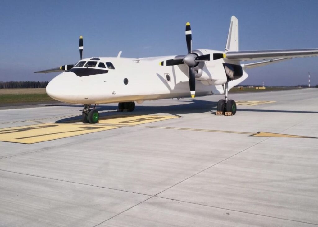 A white Antonov An-26 transport aircraft sits on a concrete runway on a clear day.