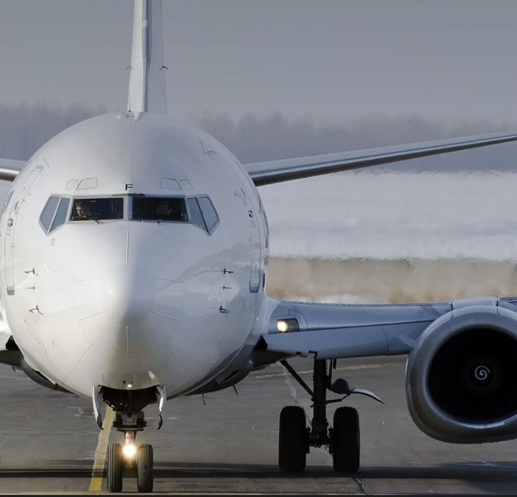 A frontal view of a white passenger airplane on the runway, with the pilots visible in the cockpit and the front landing gear and engines clearly shown.