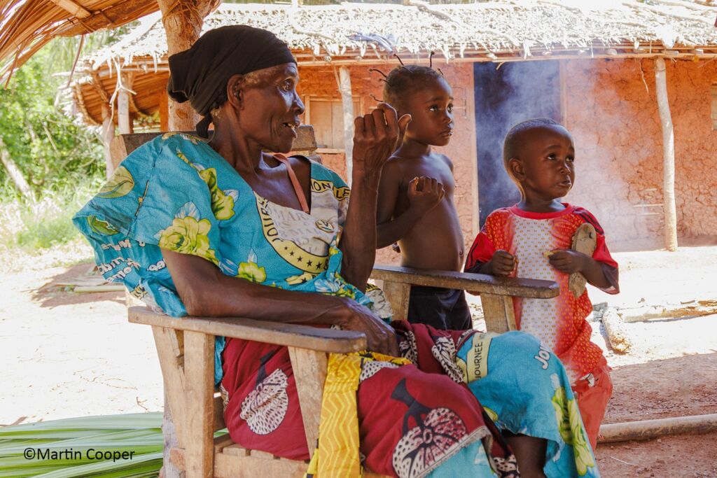 An elderly African woman sits in a wooden chair, talking, while two young children stand nearby, outside a traditional house.