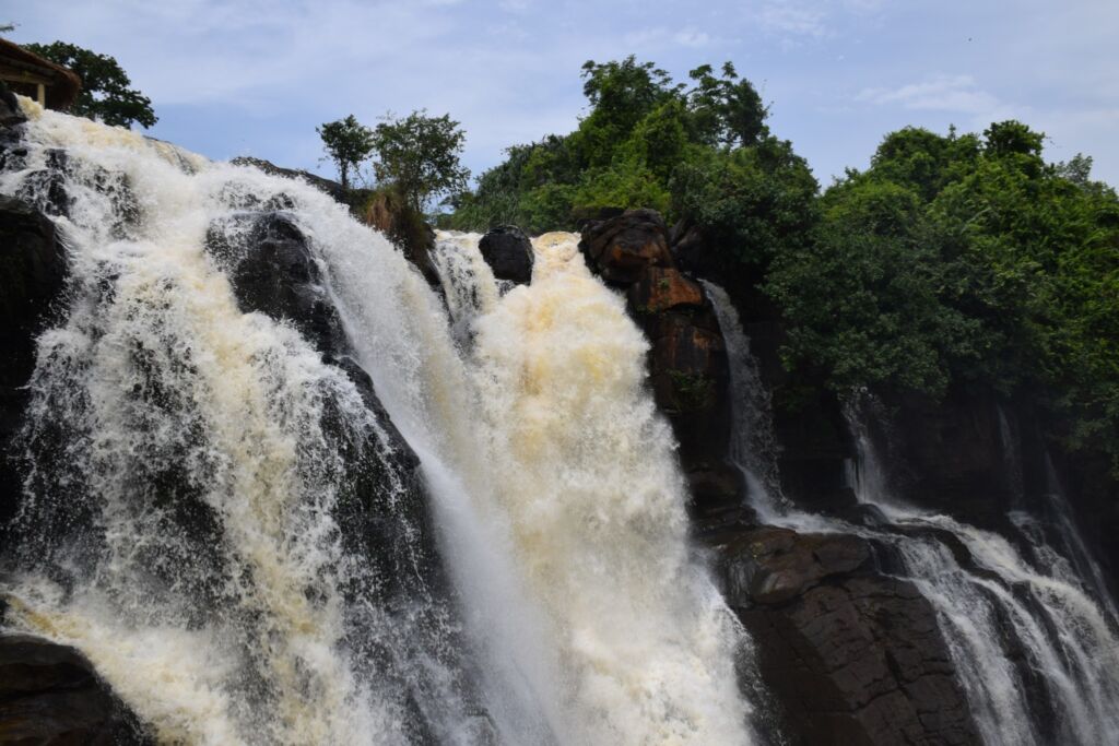 A powerful waterfall cascading over dark rocks and lush greenery under a cloudy sky.