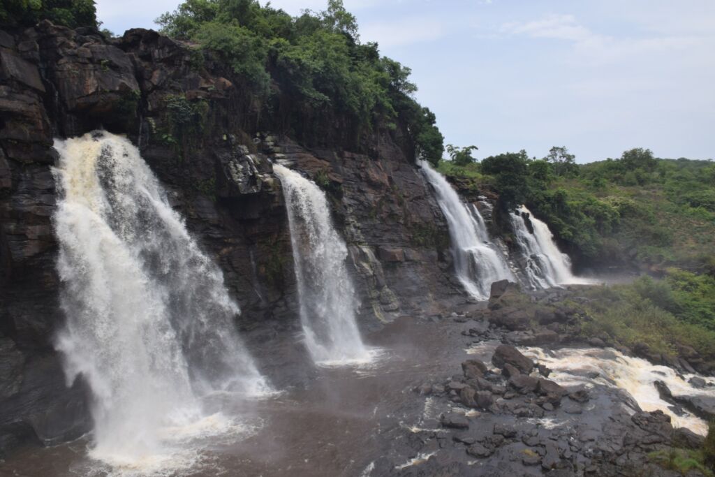 A scenic view of a rocky cliffside featuring multiple waterfalls cascading into a river, surrounded by lush green vegetation and a cloudy sky.