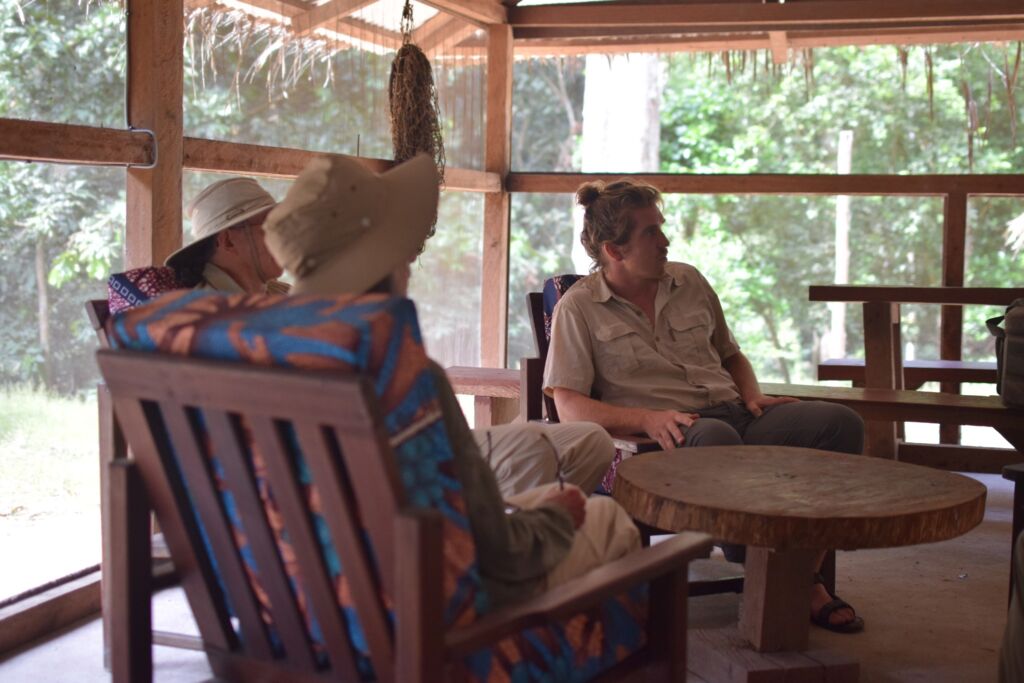 Three people are relaxing in a tropical-style outdoor lounge area with wooden furniture and lush greenery visible in the background.