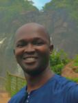 A smiling man stands in front of a waterfall in a lush green environment.