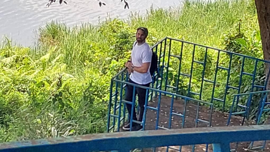 A man with a backpack stands on blue metal stairs near lush greenery, seemingly looking towards the viewer.