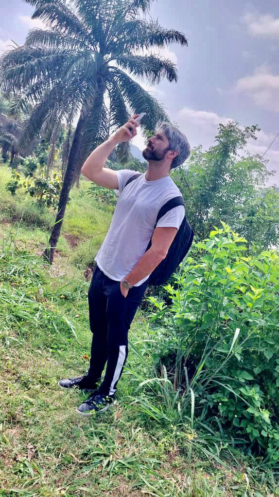 A man stands in a grassy area with a palm tree, holding a phone up to the sky, wearing a white t-shirt, black pants with a white stripe, and a backpack.
